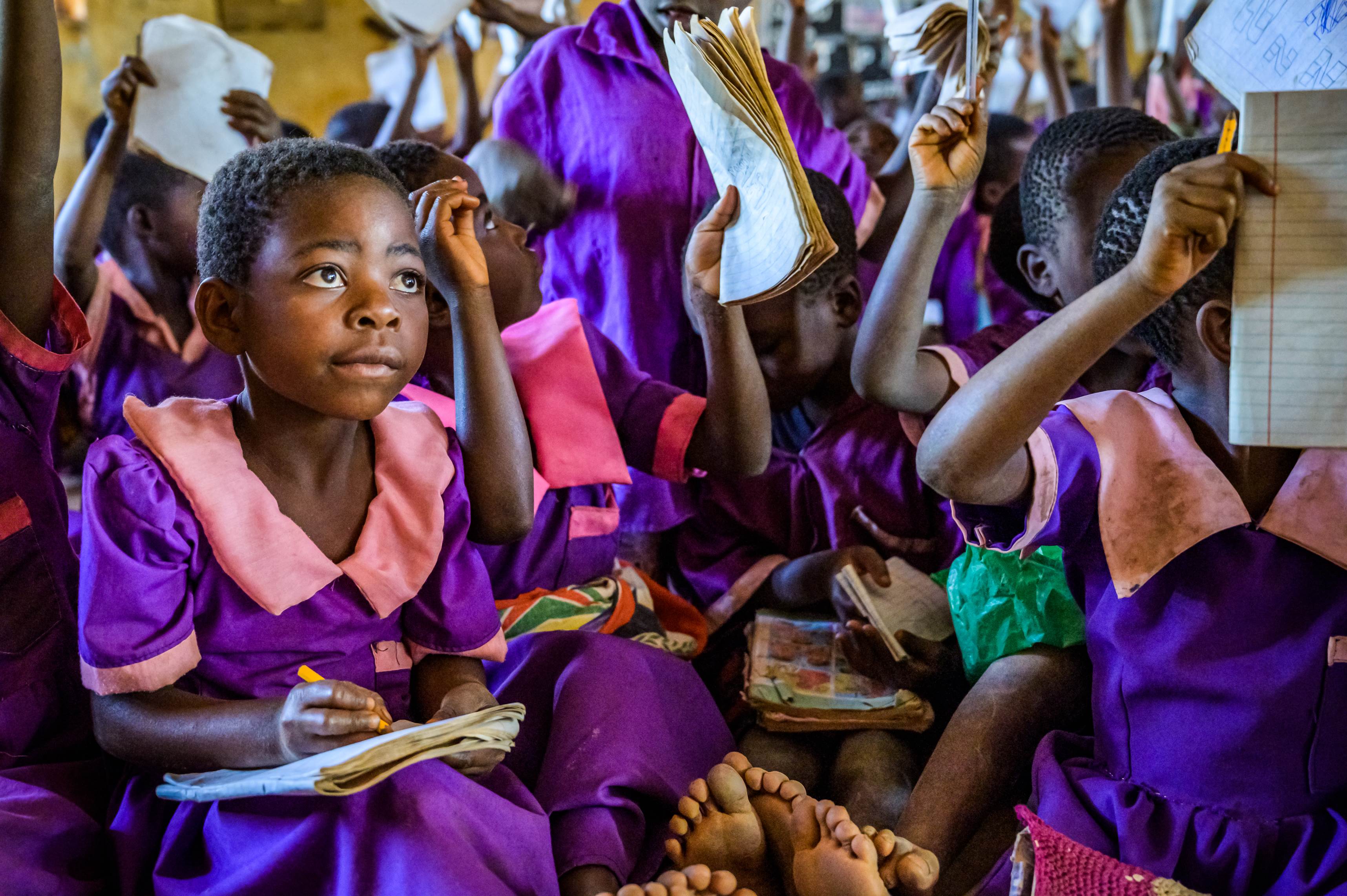 A young girl from Malawi sat in a classroom with a workbook and listening, with lots of other children in the background