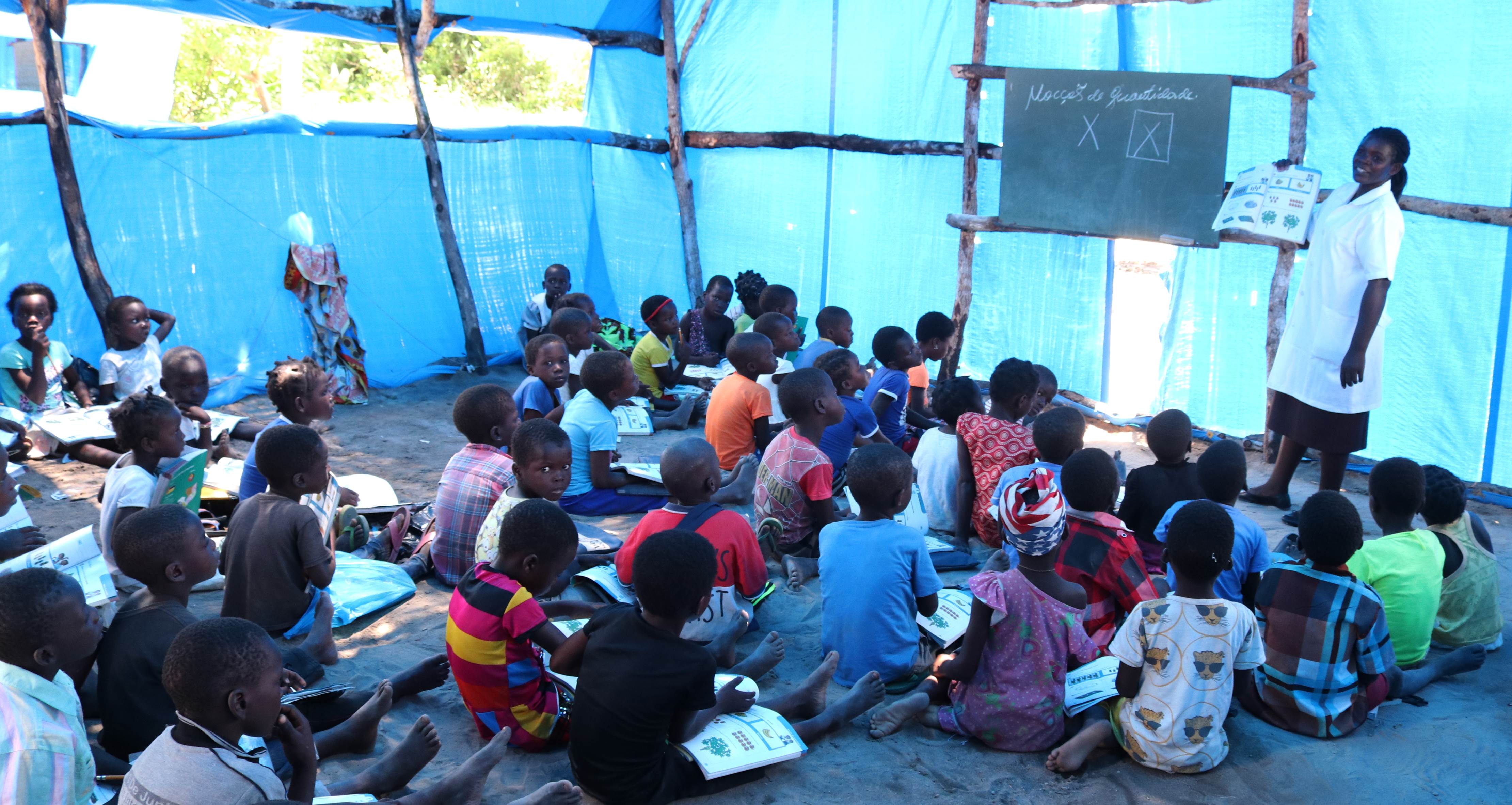 A blue tent in Mozambique is full of people sitting and looking at a makeshift board for teaching