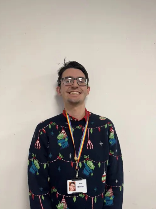 Sam grins in a photo. He's wearing a blue Christmas jumper.