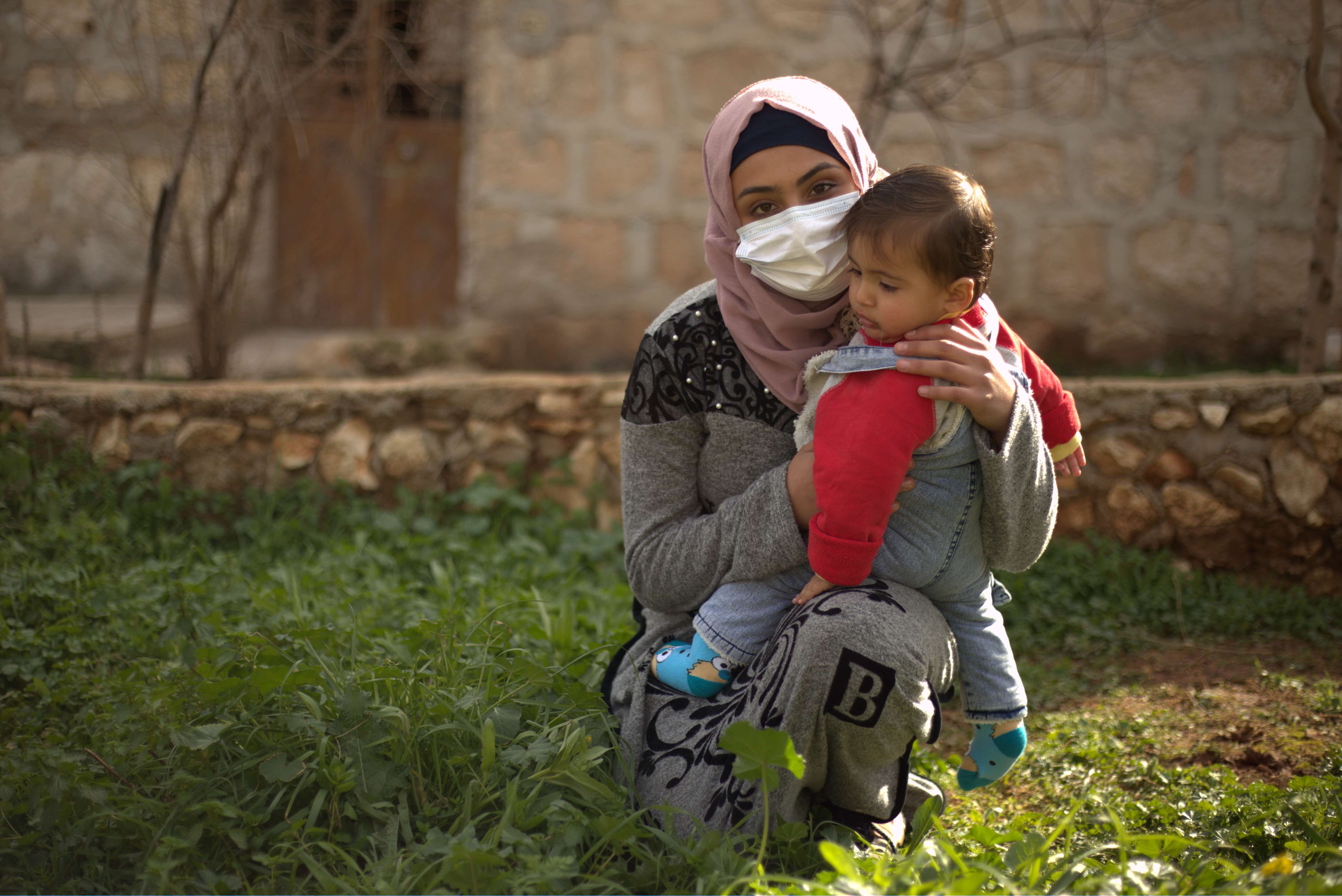 A Syrian refugee woman with a mask holds her toddler outside in Jordan