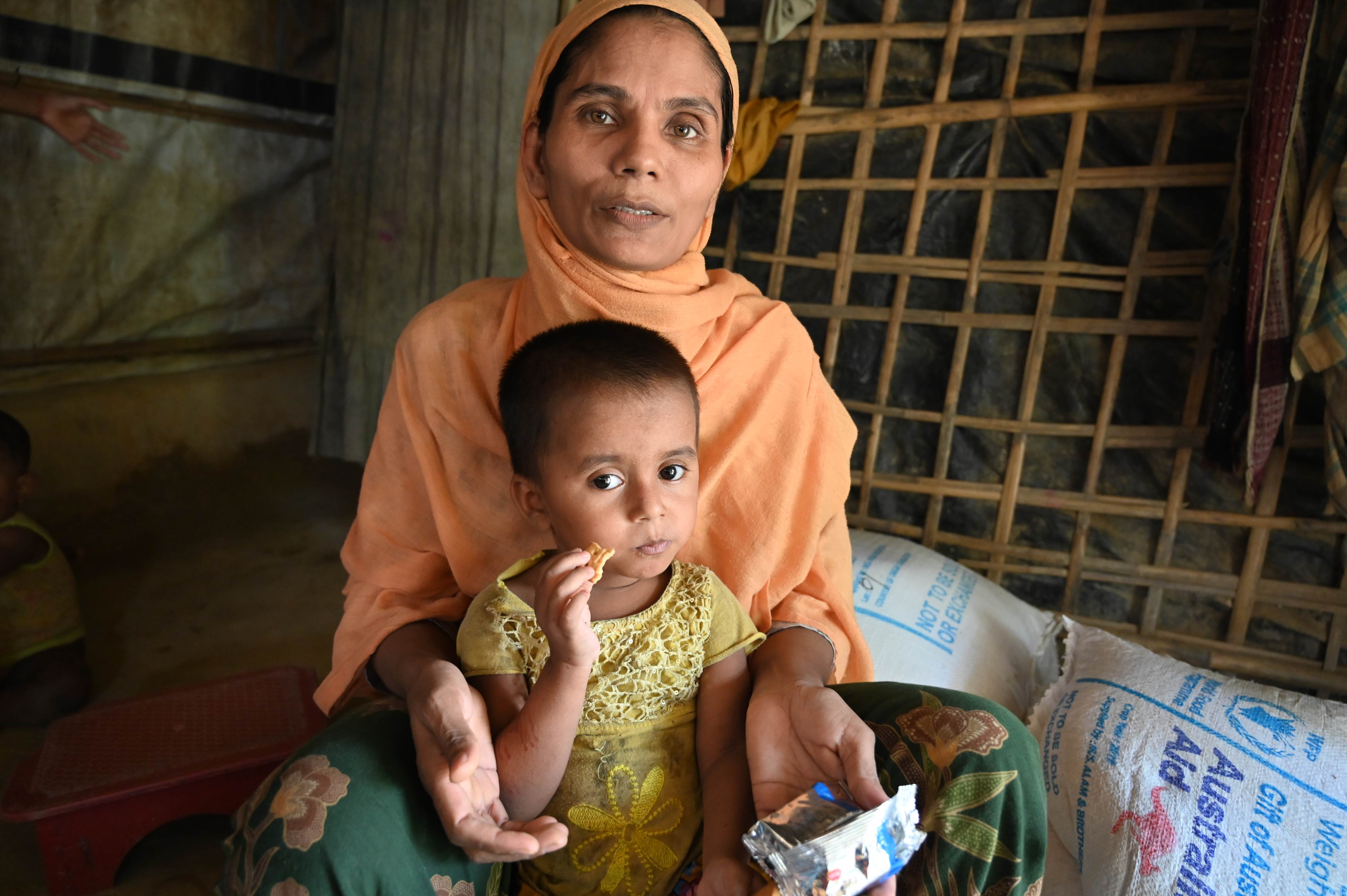 Mother sits with her child on her lap in a dark room, the child eating World Vision provisions