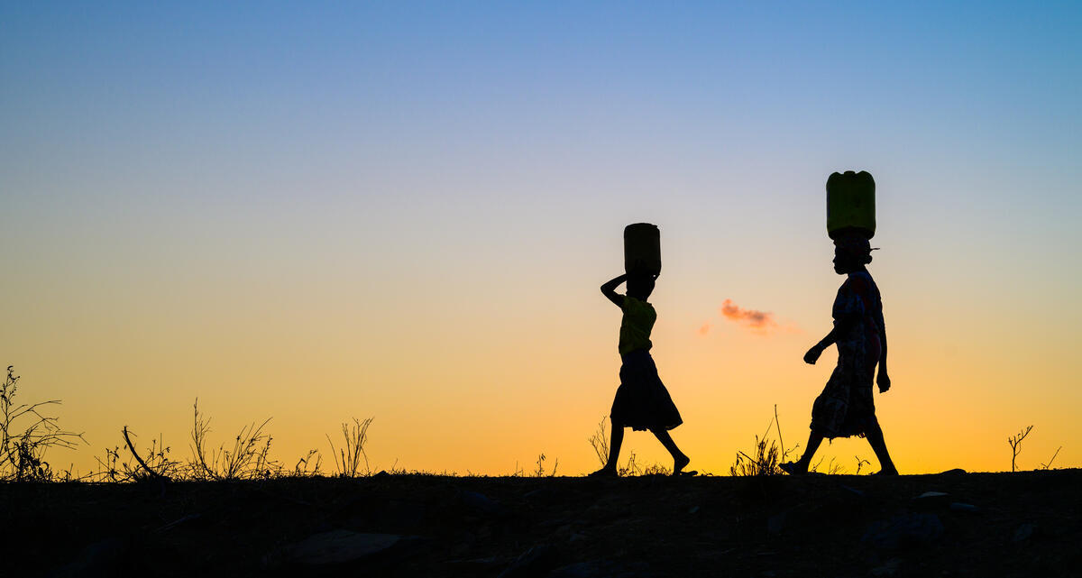 A silhouette image of a girl and a woman walking in the dusk with water cans on their heads