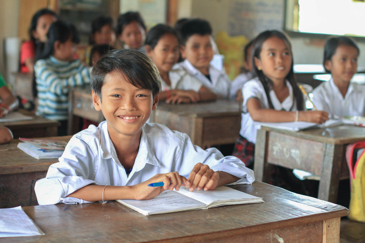 Child in Cambodia sits at their desk in a busy classroom, hands resting over their workbook and smiling at the camera