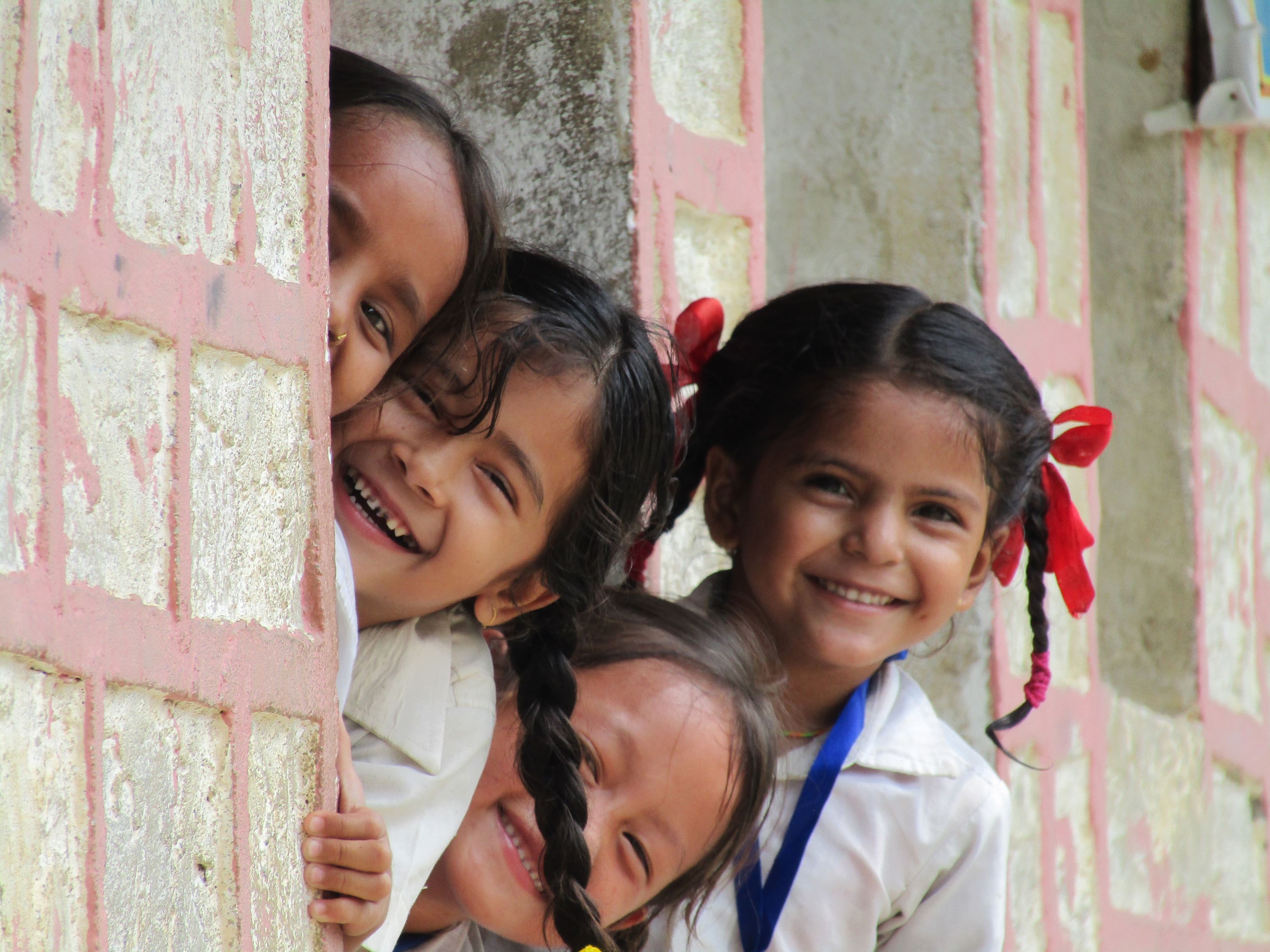 4 Nepalese children peek round a wall, smiling and giggling