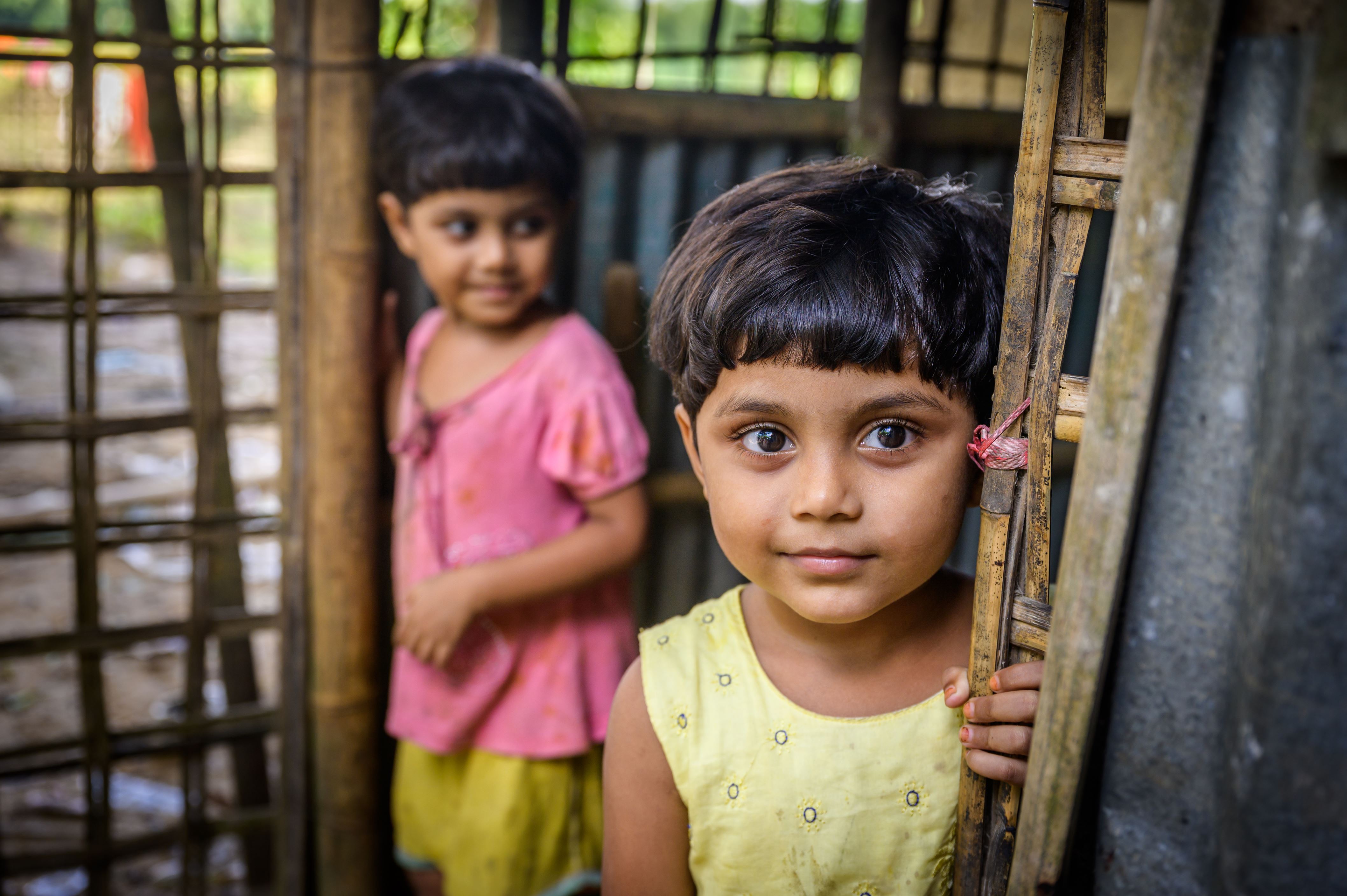 Twin girls from Bangladesh stand at the doorway from their house, one in yellow, one in pink