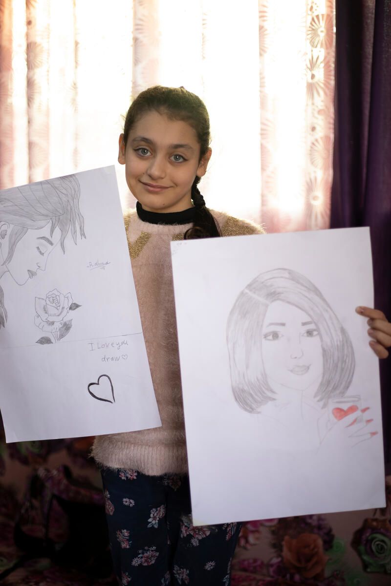 A young girl holding and displaying her drawings, which helps her escape from the worries of war