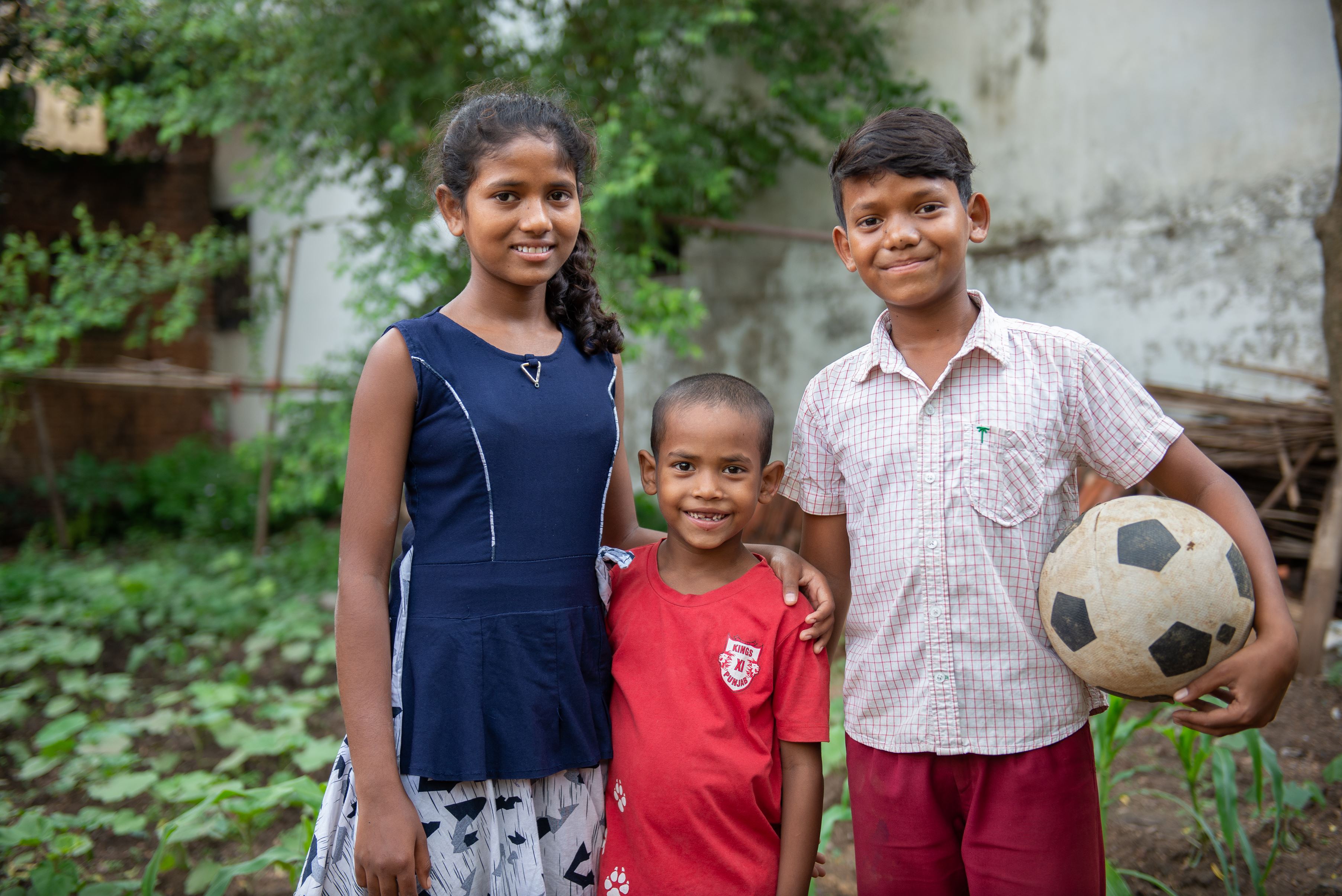 Three children from India smile into the camera, one boy holding a football