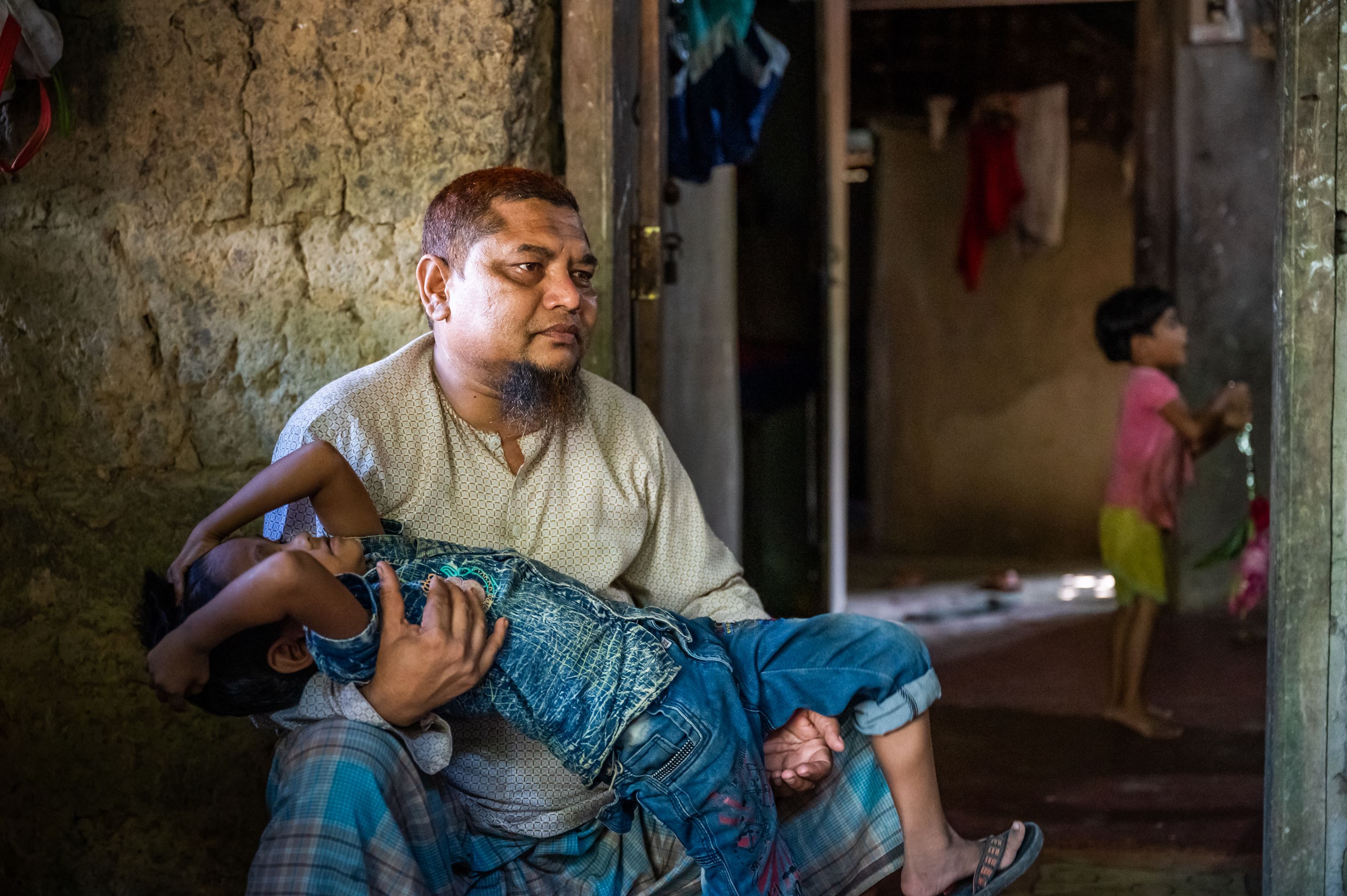 Man holds his son in his arms as he looks away from the camera, sitting in his house in Bangladesh