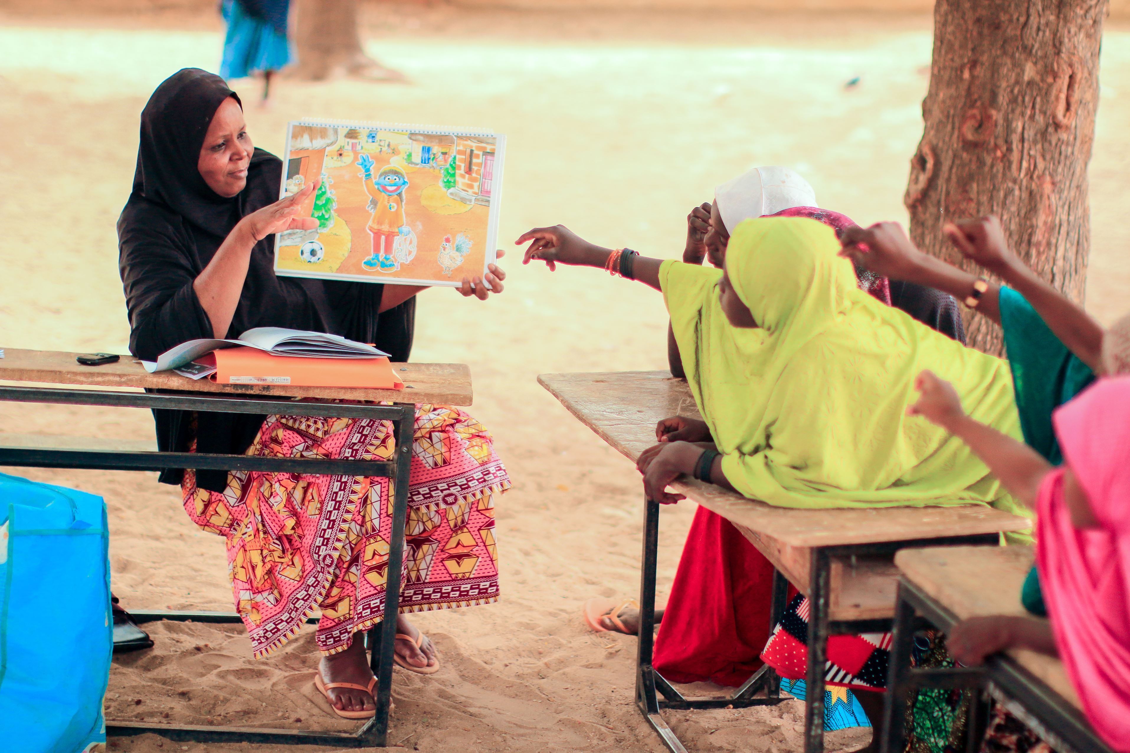 Woman from Niger sits outside with a group of children at desks, teaching