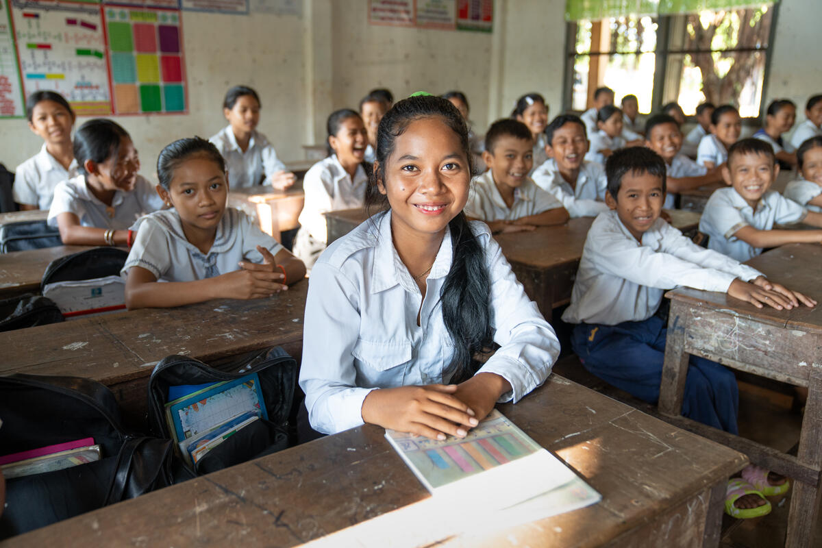 A young girl from Cambodia sat in a classroom with a workbook and listening, with lots of other children in the background