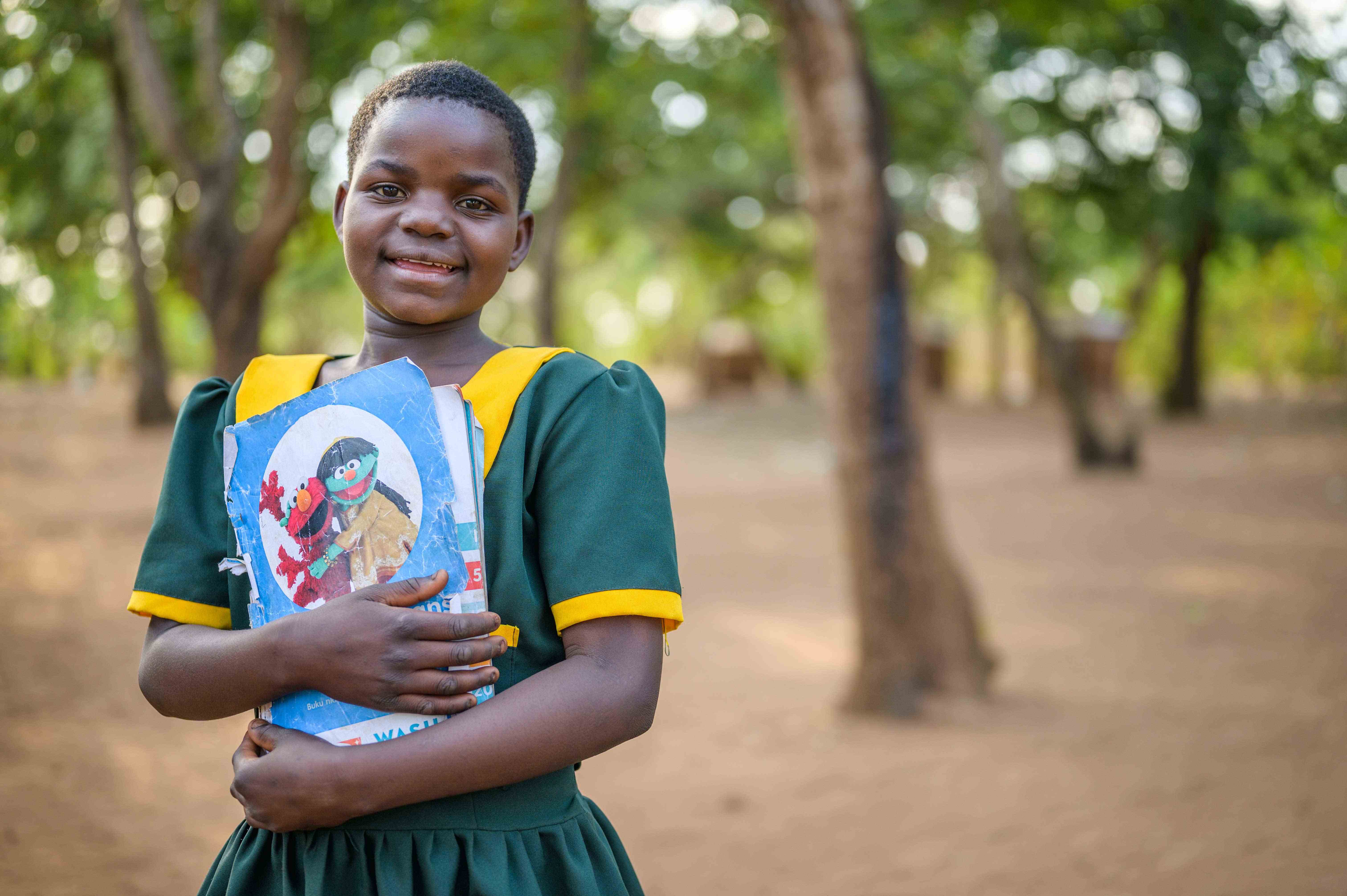 Girl from Malawi grins and holds her book, standing outside in front of trees