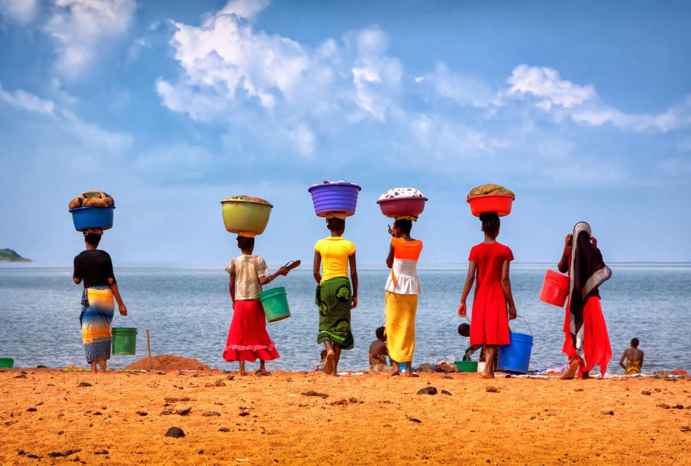 Six Tanzanian women walking away from the camera, towards the ocean with buckets balanced on their heads