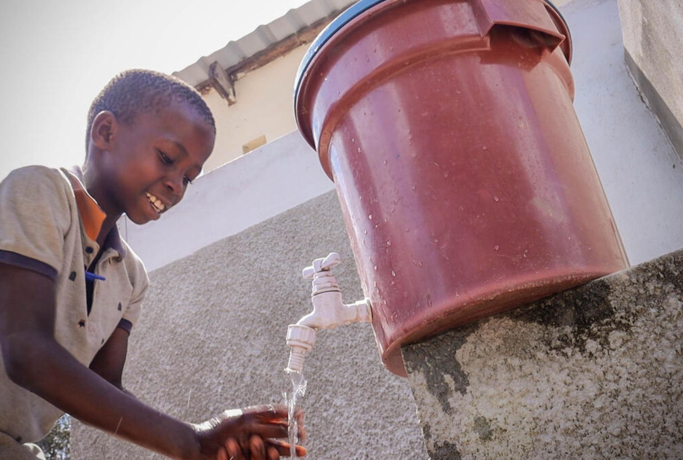 Child in Mozambique smiles as he washes his hands with water dispensing from a bucket with a tap