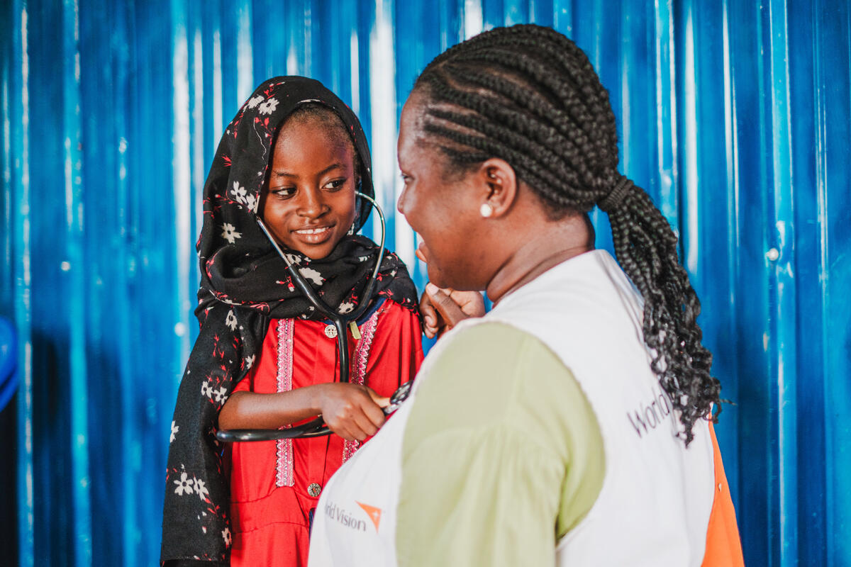 A young girl holding a stethoscope to a World Vision staff member and is smiling