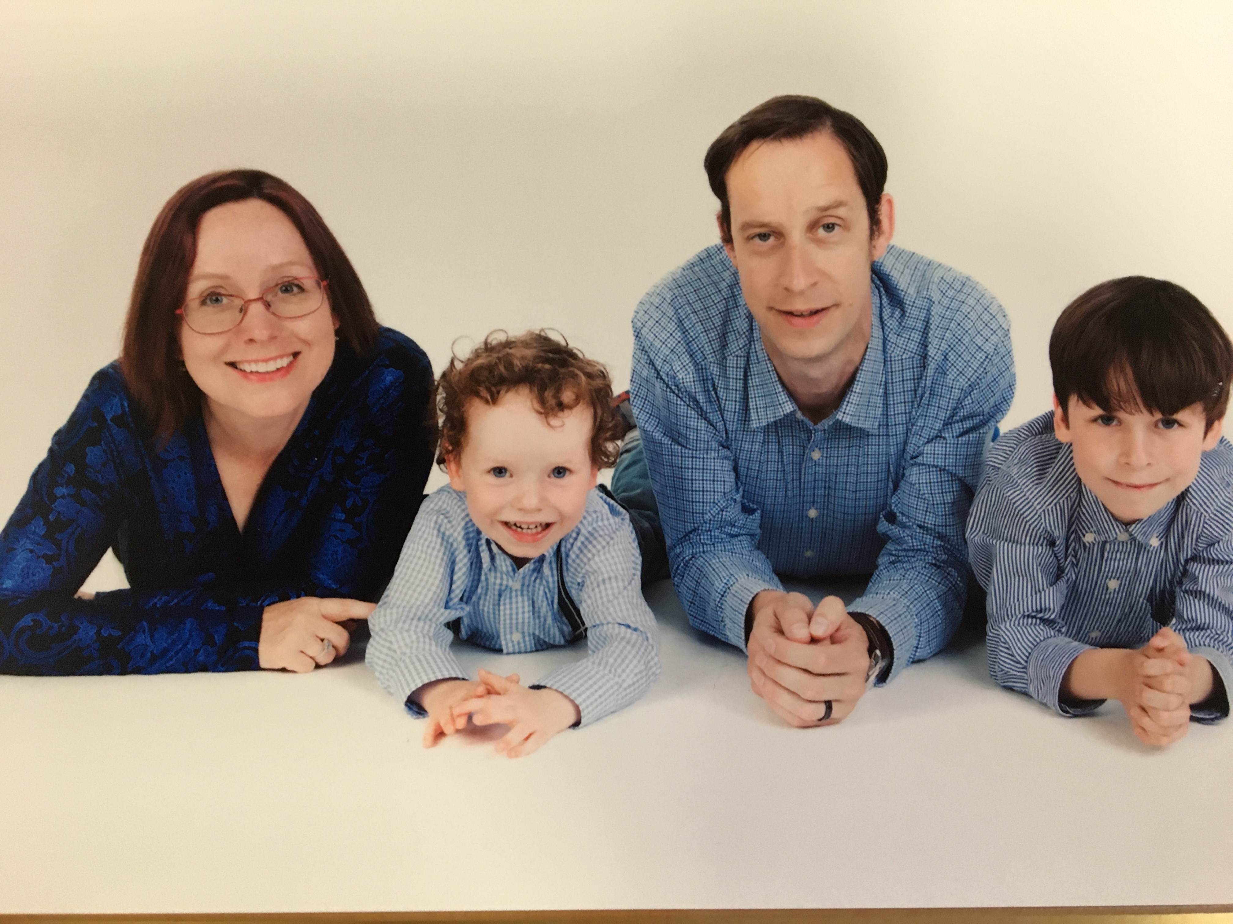 Two adults and two children dressed in blue tops lean against a white table with a white backdrop