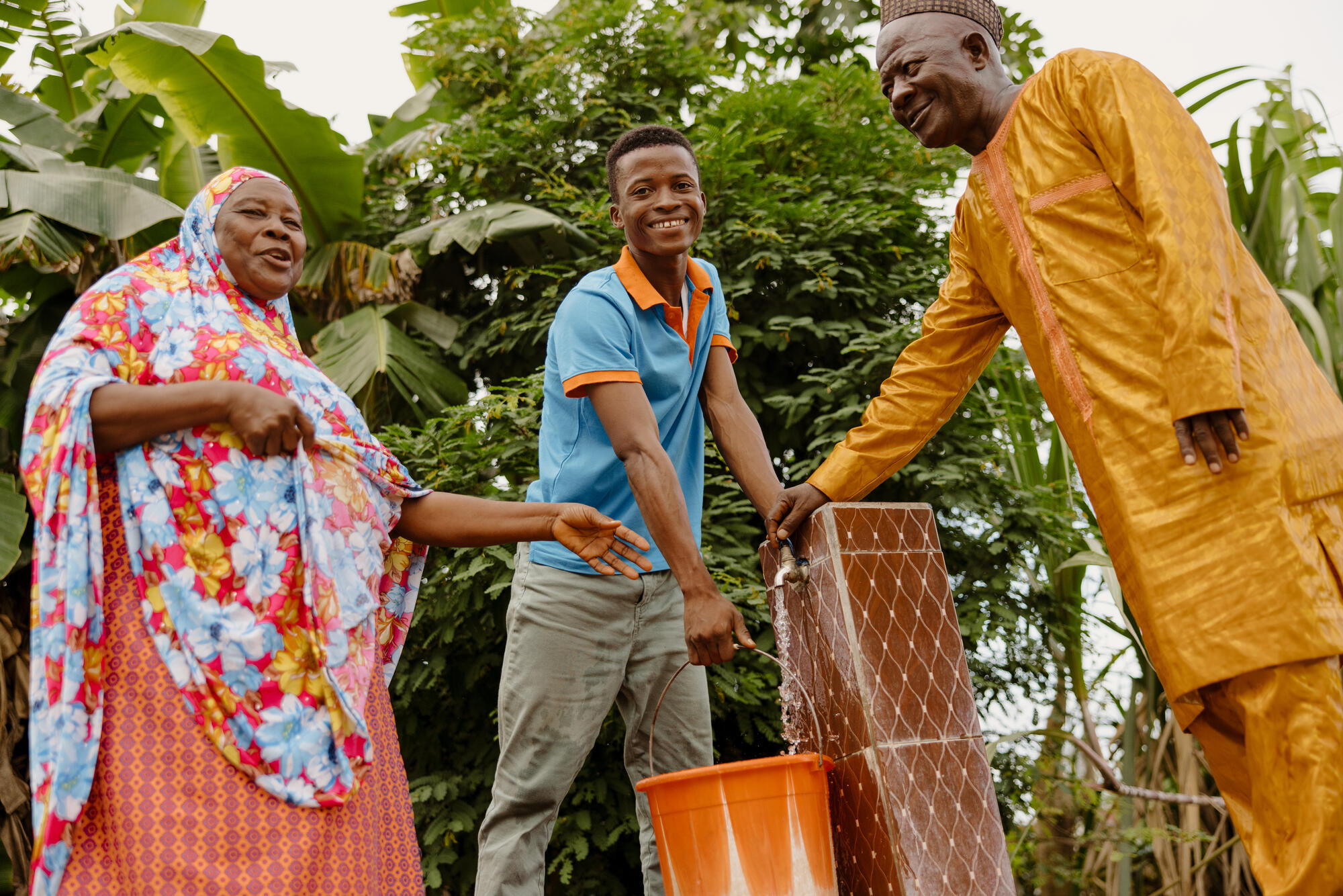 Three adults pictured smiling next to water tap, Sierra Leone.