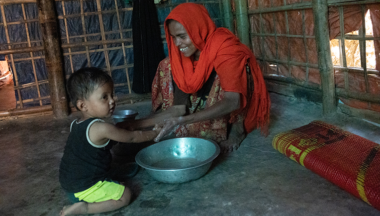 Woman in Bangladesh sits in a room with wooden walls, cleaning the hands of a child using a metal pot of water