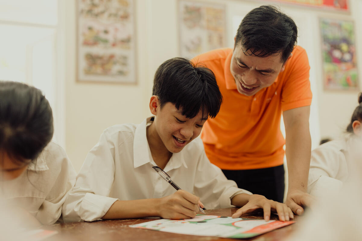 A boy in Vietnam writes to their sponsor, sat alongside a World Vision staff member