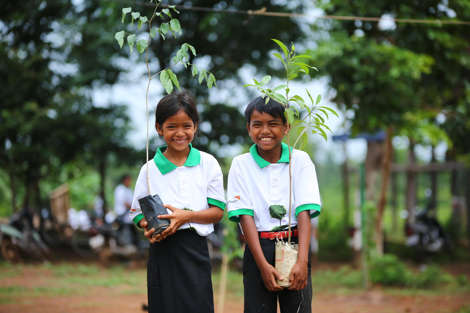 Two Cambodian children smile while holding small trees before planting them.