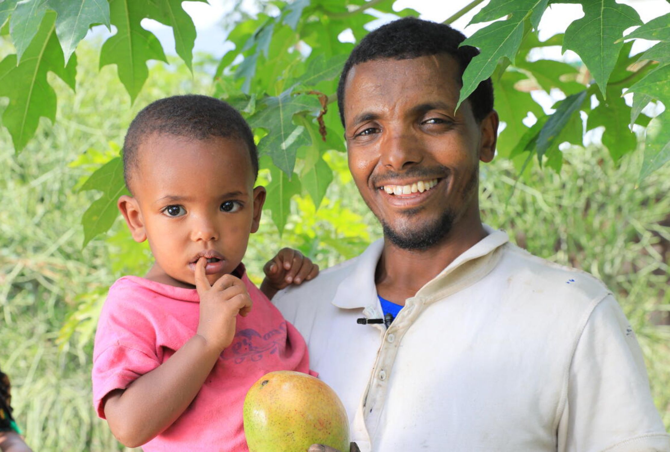 Ethiopian dad smiling at the camera, holding his child and a ripe mango