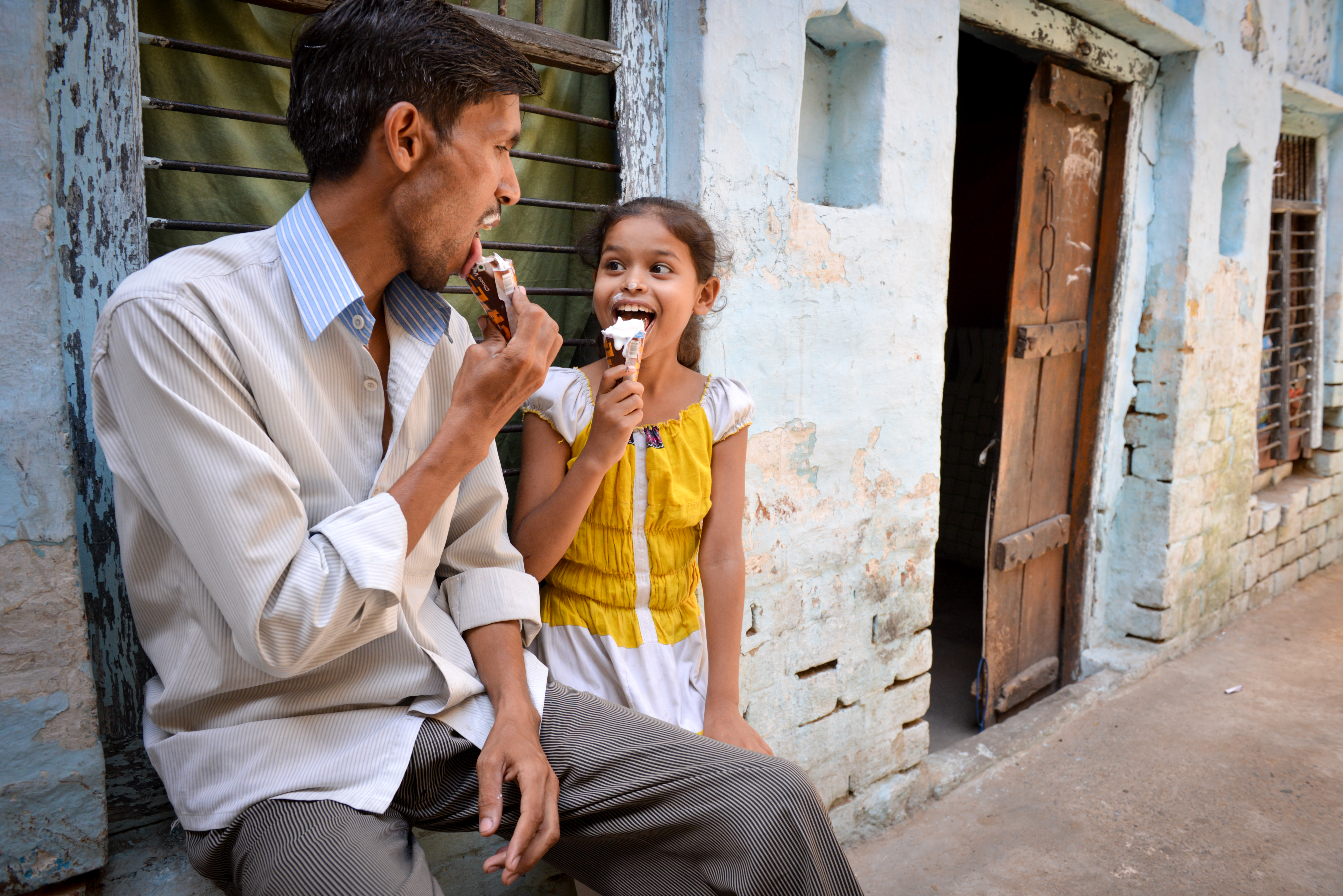 A young girl and her dad perch together on a outside windowsill  -each enjoying an ice cream