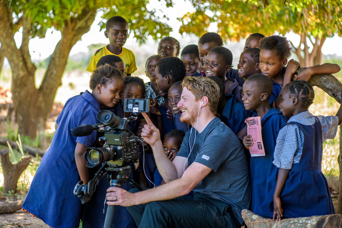World Vision videographer with crowd of young children in Zambia smile around a camera
